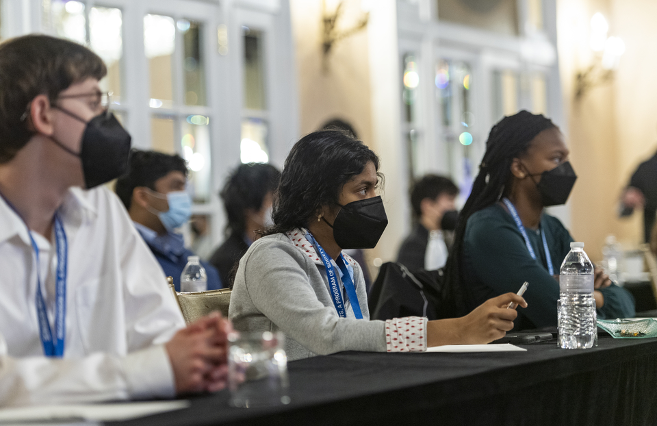 在STS决赛周期间，三名戴着口罩的Regeneron Science Scient Search搜索决赛选手戴着口罩坐在面对扬声器的桌子上