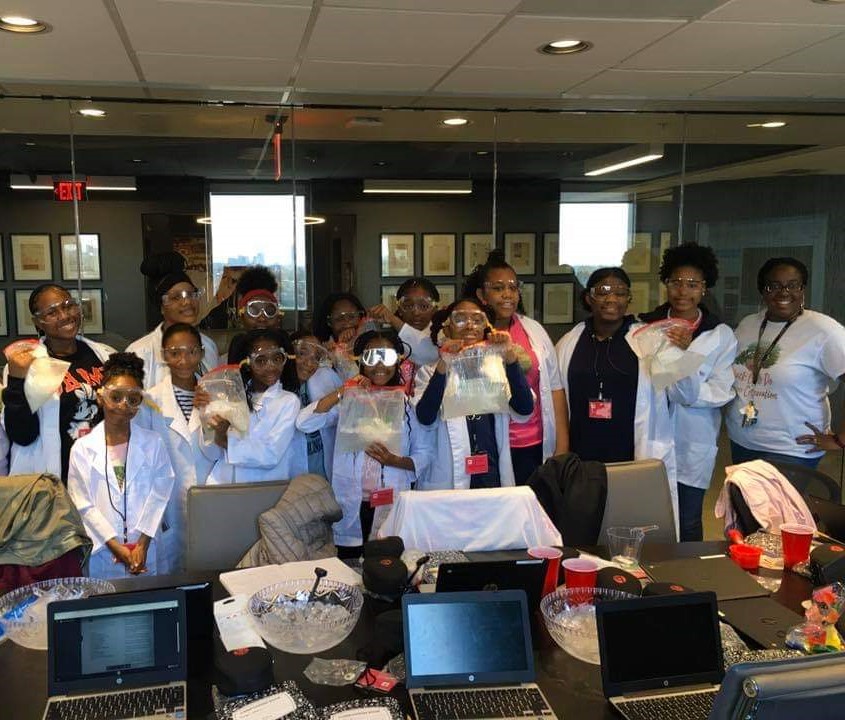 一群年轻女孩聚集在一起，穿着白色实验室外套和护目镜。